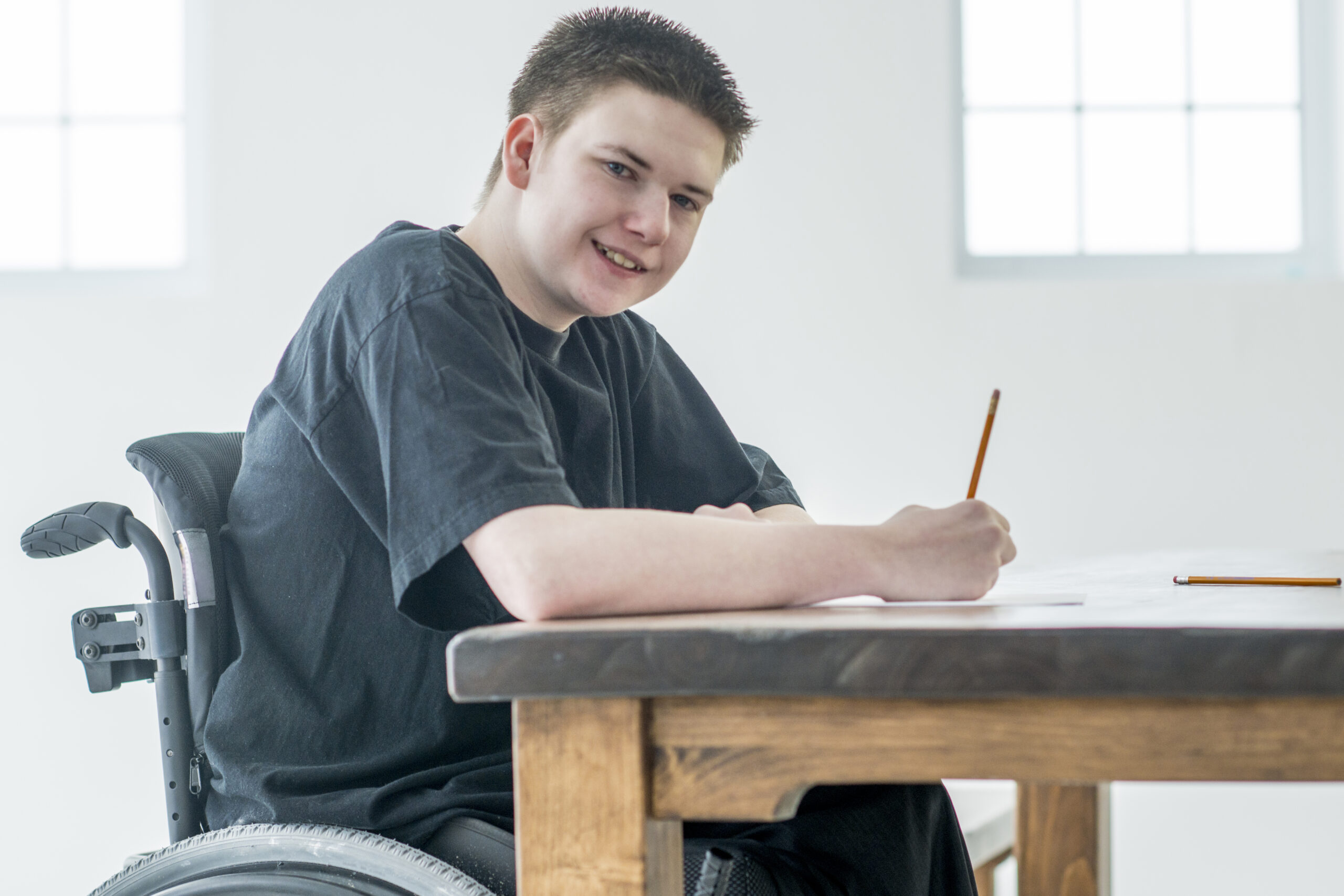 A young boy with a physical disability is working on a homework assignment from home. He is smiling and looking at the camera.